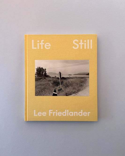 A hardcover book titled 'Life Still' by Lee Friedlander, featuring a mustard yellow cover with a central black-and-white landscape photograph, photographed against a neutral light gray background.